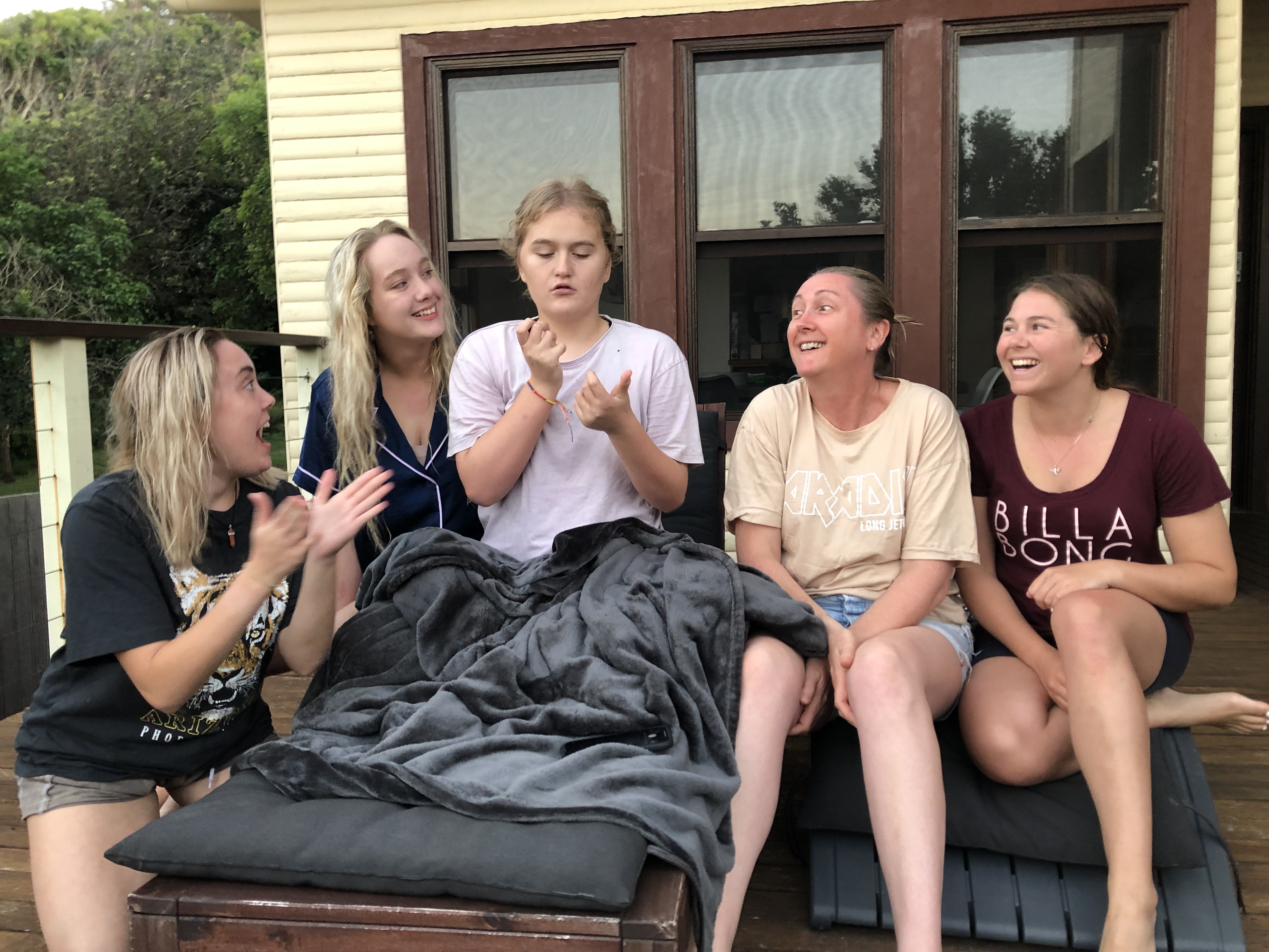 Abbey is surrounded by her team of support workers and her mum. They're sitting on the balcony of their home. Abbey is gesturing with her hands while her support workers look to her. 