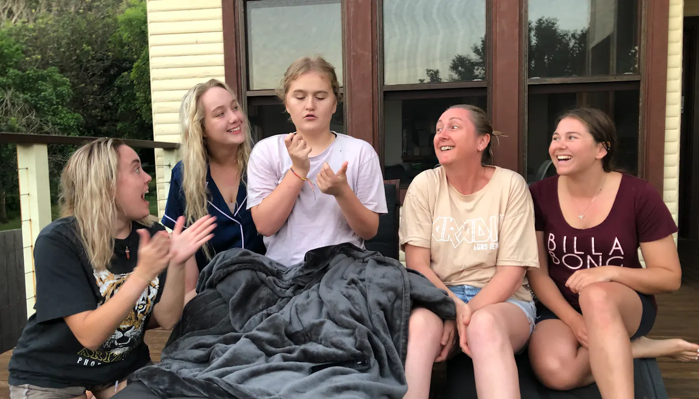 Abbey is surrounded by her team of support workers and her mum. They're sitting on the balcony of their home. Abbey is gesturing with her hands while her support workers look to her.