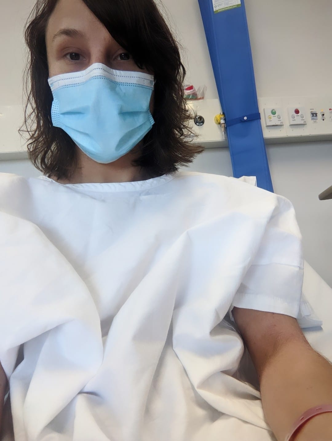 Elena is sitting up in a hospital bed. She has dark shoulder length hair and is wearing a white hospital gown and a blue facemask. 