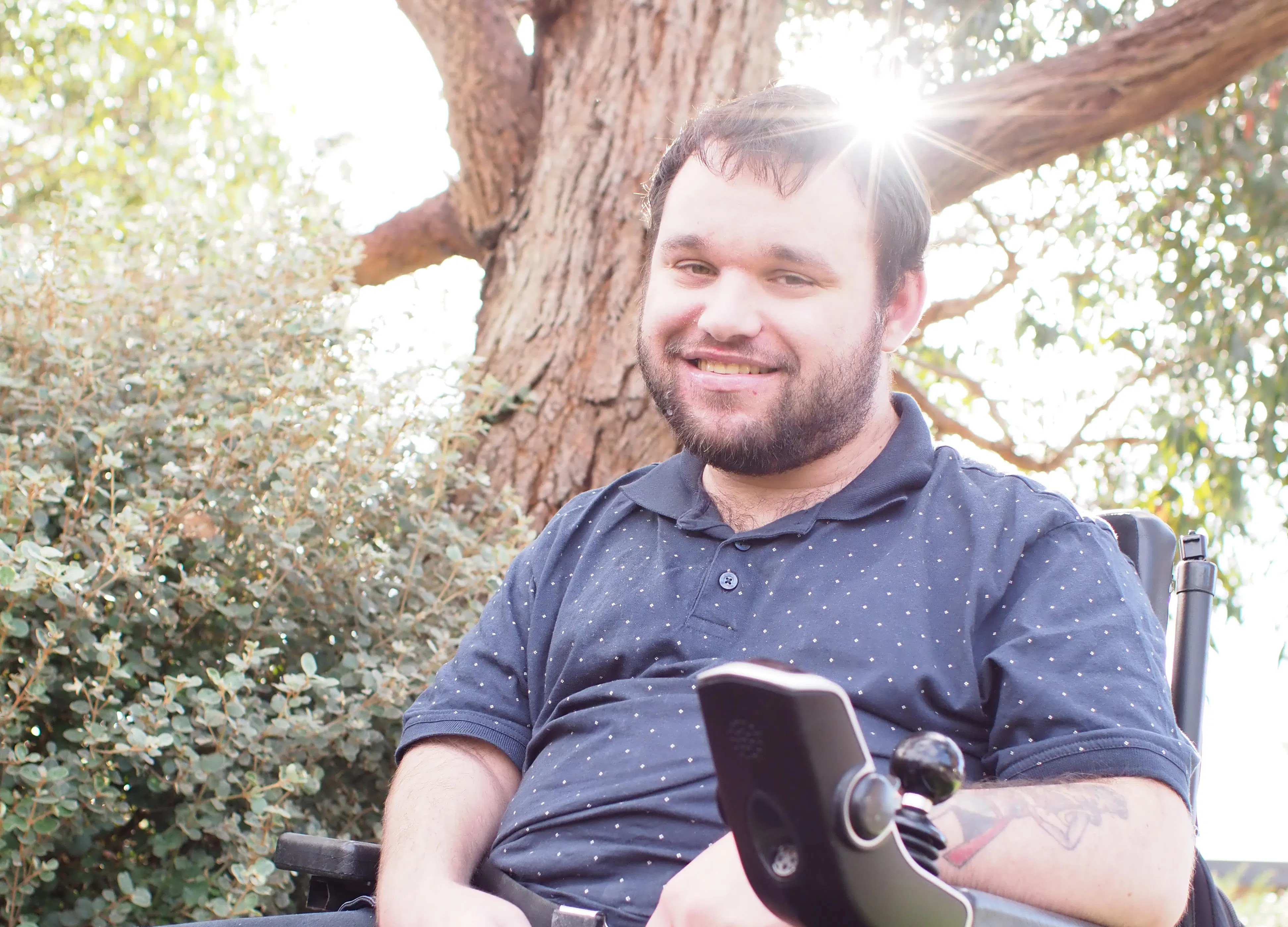 Jonathan has a trimmed beard and is smiling. There is sun shining through a tree in the background. He is wearing a blue shirt. Jonathan uses a wheelchair.