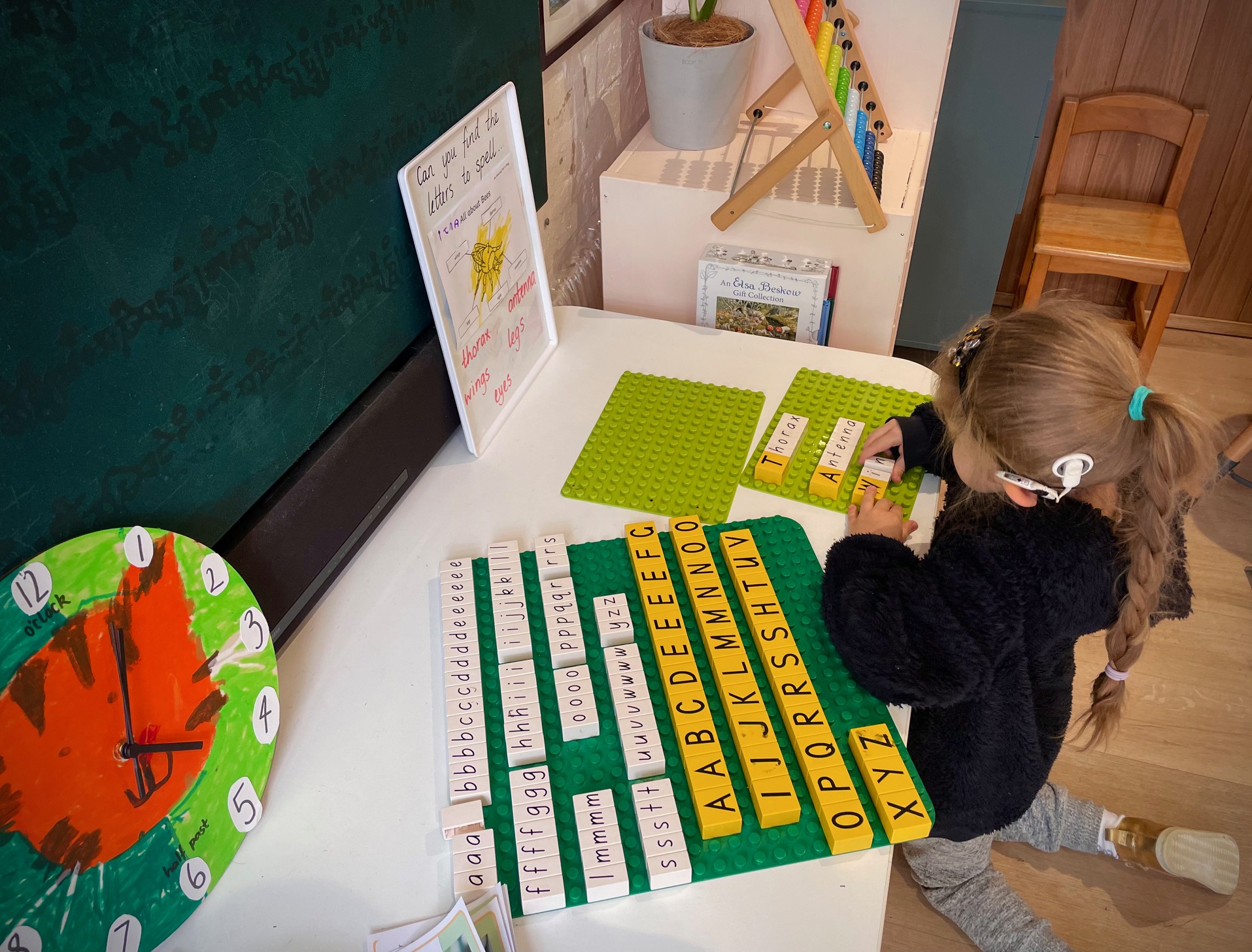 A young girl with a hearing aid spells out words with plastic letter tiles on a green board. She kneels in a room filled with educational toys and tools. 