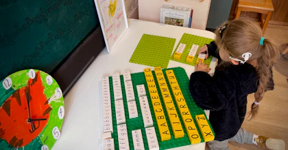 A young girl with a hearing aid spells out words with plastic letter tiles on a green board. She kneels in a room filled with educational toys and tools.
