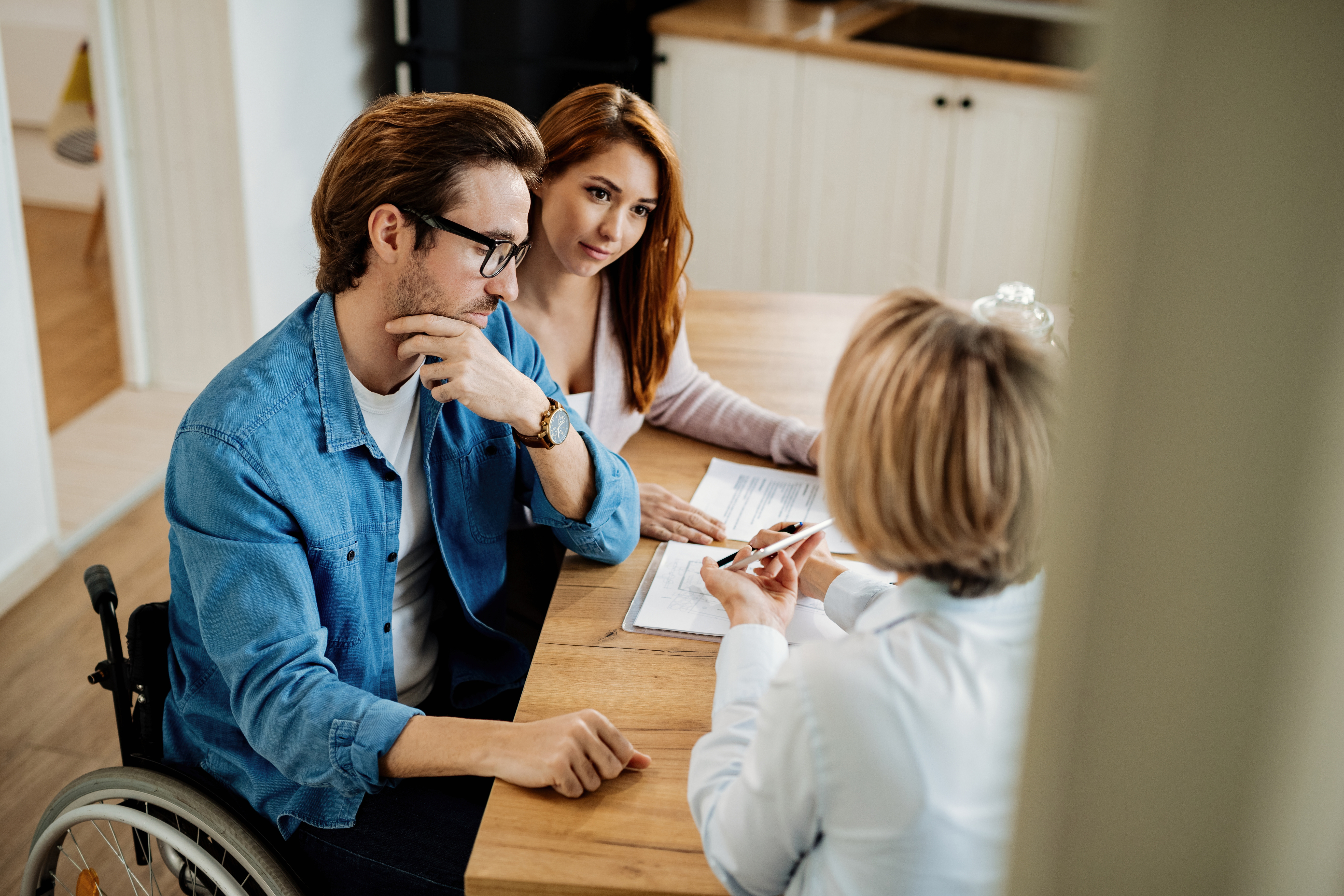 A man, woman and two women are sitting at a table looking at some paperwork. The man has short brown hair, wears black glasses and is a wheelchair user. 