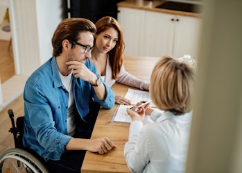 A man, woman and two women are sitting at a table looking at some paperwork. The man has short brown hair, wears black glasses and is a wheelchair user.