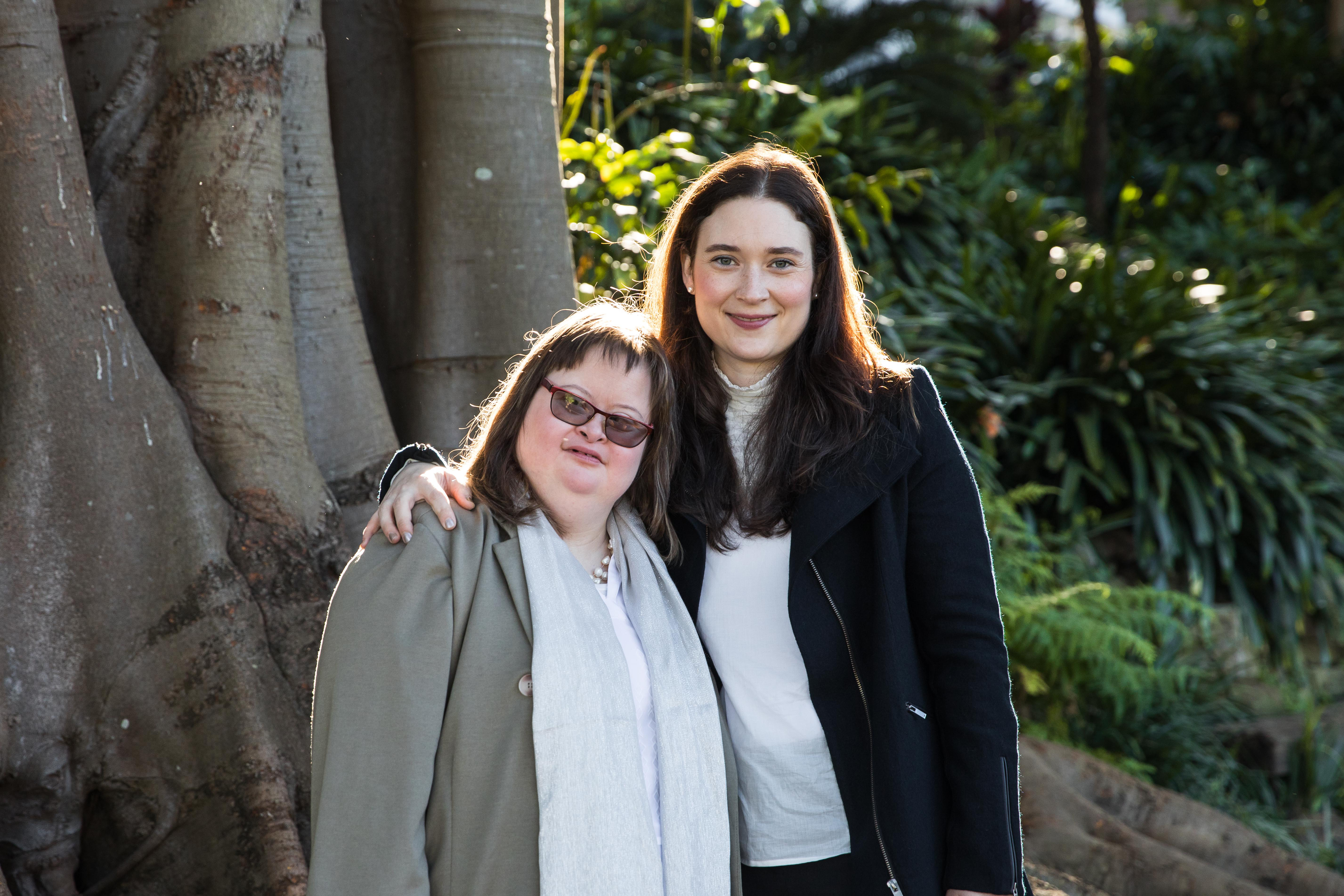 Hannah is standing in front of a tree with her sister Camilla. She has her arm around Camilla's shoulder. Camilla and Hannah are smiling at the camera. 