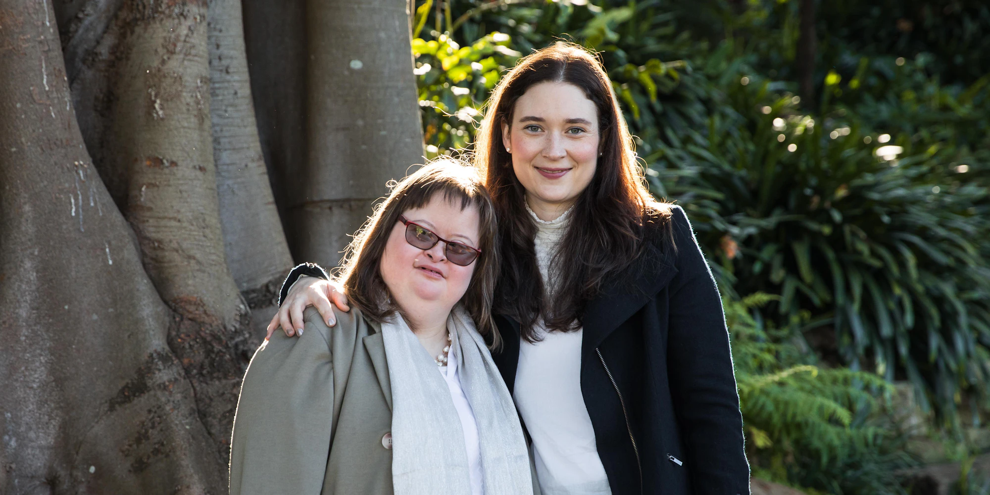 Hannah is standing in front of a tree with her sister Camilla. She has her arm around Camilla's shoulder. Camilla and Hannah are smiling at the camera.
