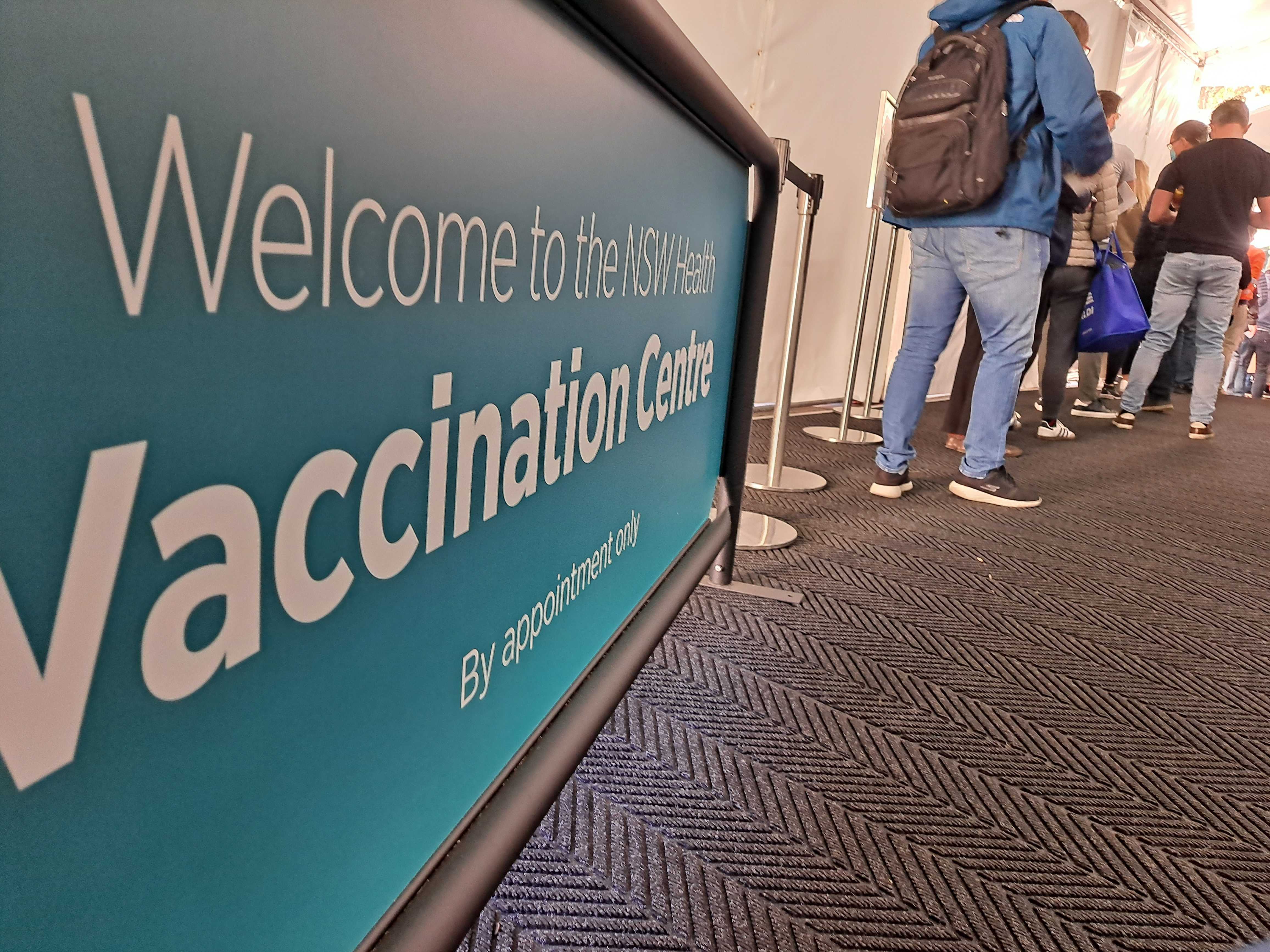 A blue and white banner reads: 'Welcome to the NSW Health Vaccination Centre. By appointment only'. A queue of people are standing near the sign. 