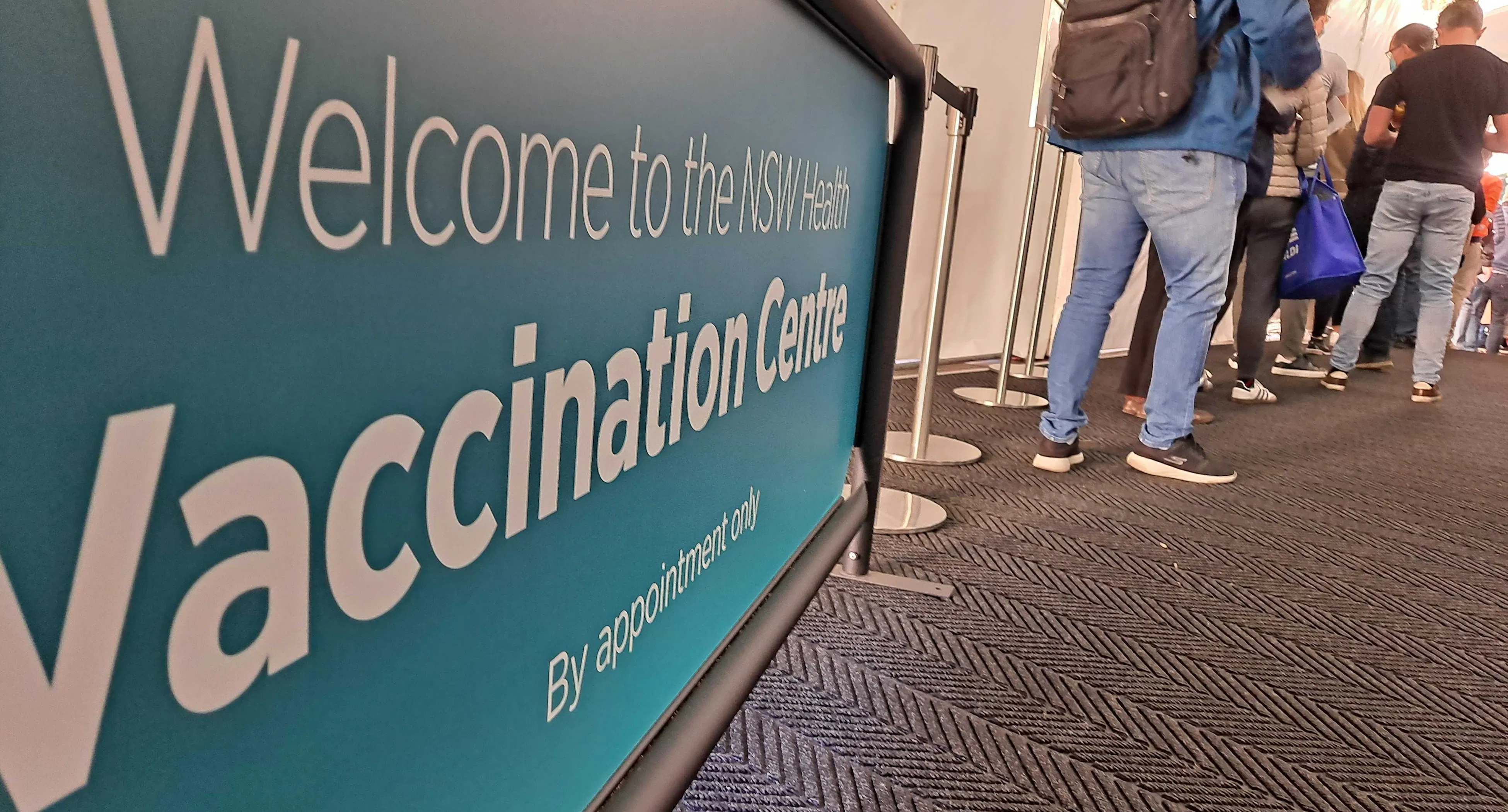 A blue and white banner reads: 'Welcome to the NSW Health Vaccination Centre. By appointment only'. A queue of people are standing near the sign.