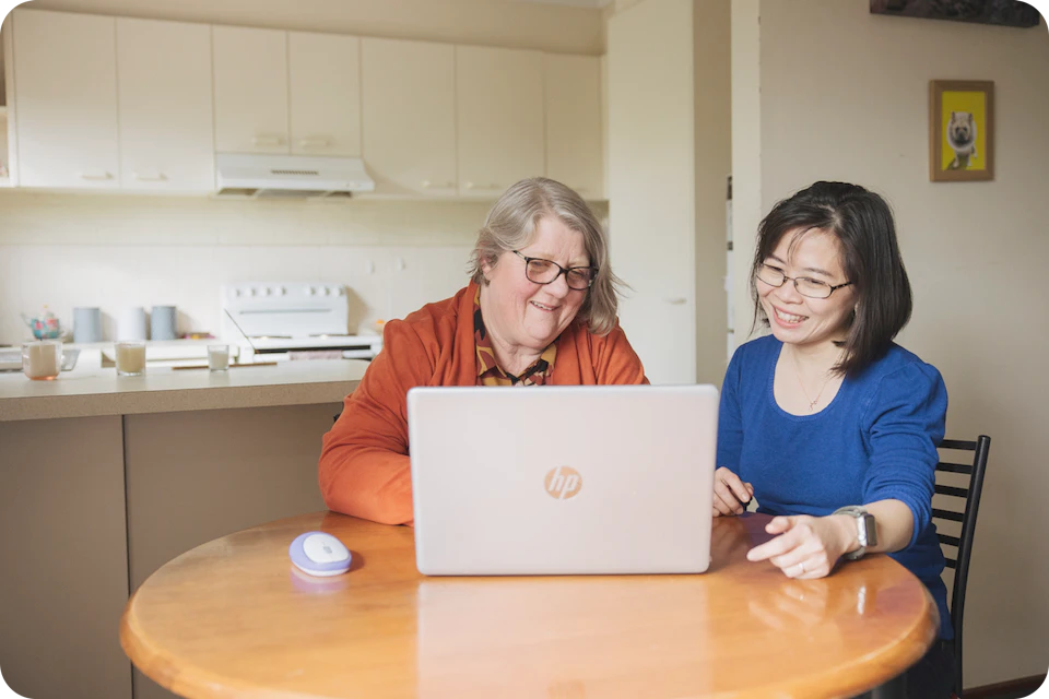 Hireup client Leanne and her support worker Dieu are seated in front of a laptop screen. They're both looking at the laptop and are smiling.