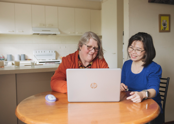 Hireup client Leanne and her support worker Dieu are seated in front of a laptop screen. They're both looking at the laptop and are smiling.