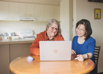 Hireup client Leanne and her support worker Dieu are seated in front of a laptop screen. They're both looking at the laptop and are smiling.