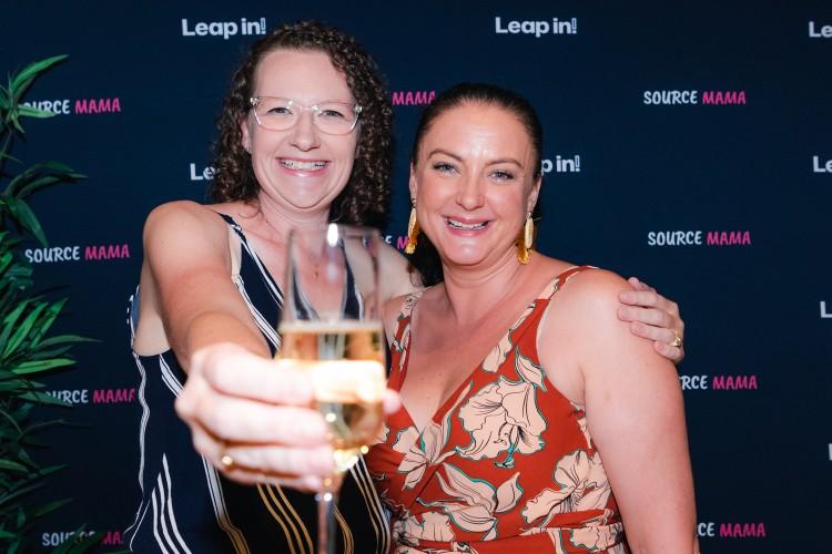 Stacey has curly hair and is holding a champagne glass, as if toasting the photographer. Sarah is wearing long earrings and a summer dress. They are standing is front of a board which has the word "Source Mama" written on it multiple times. 