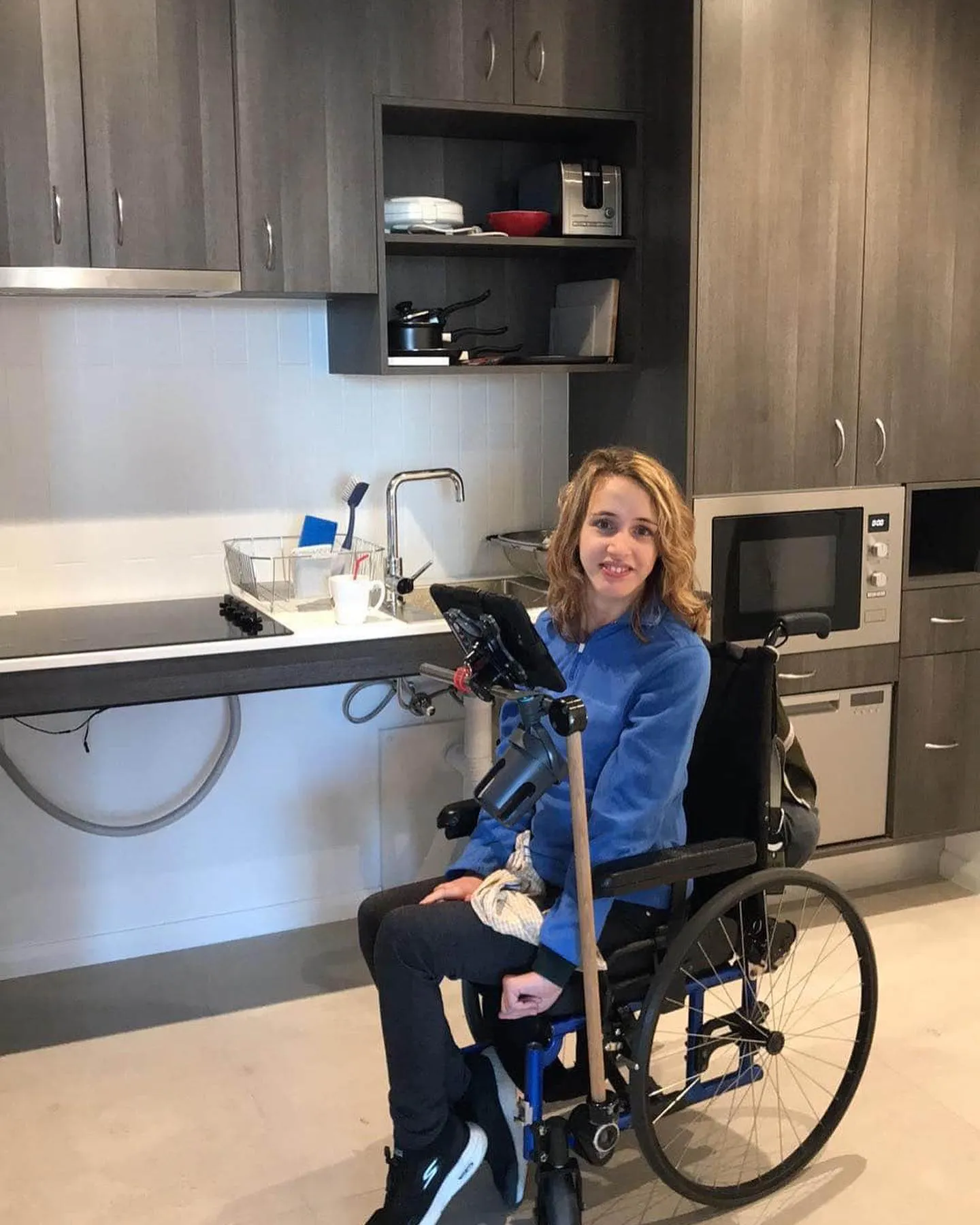 Gretta is in front of her new kitchen. There is a sink and microwave in the background. Gretta is seated on her chair with her communication device in front of her.