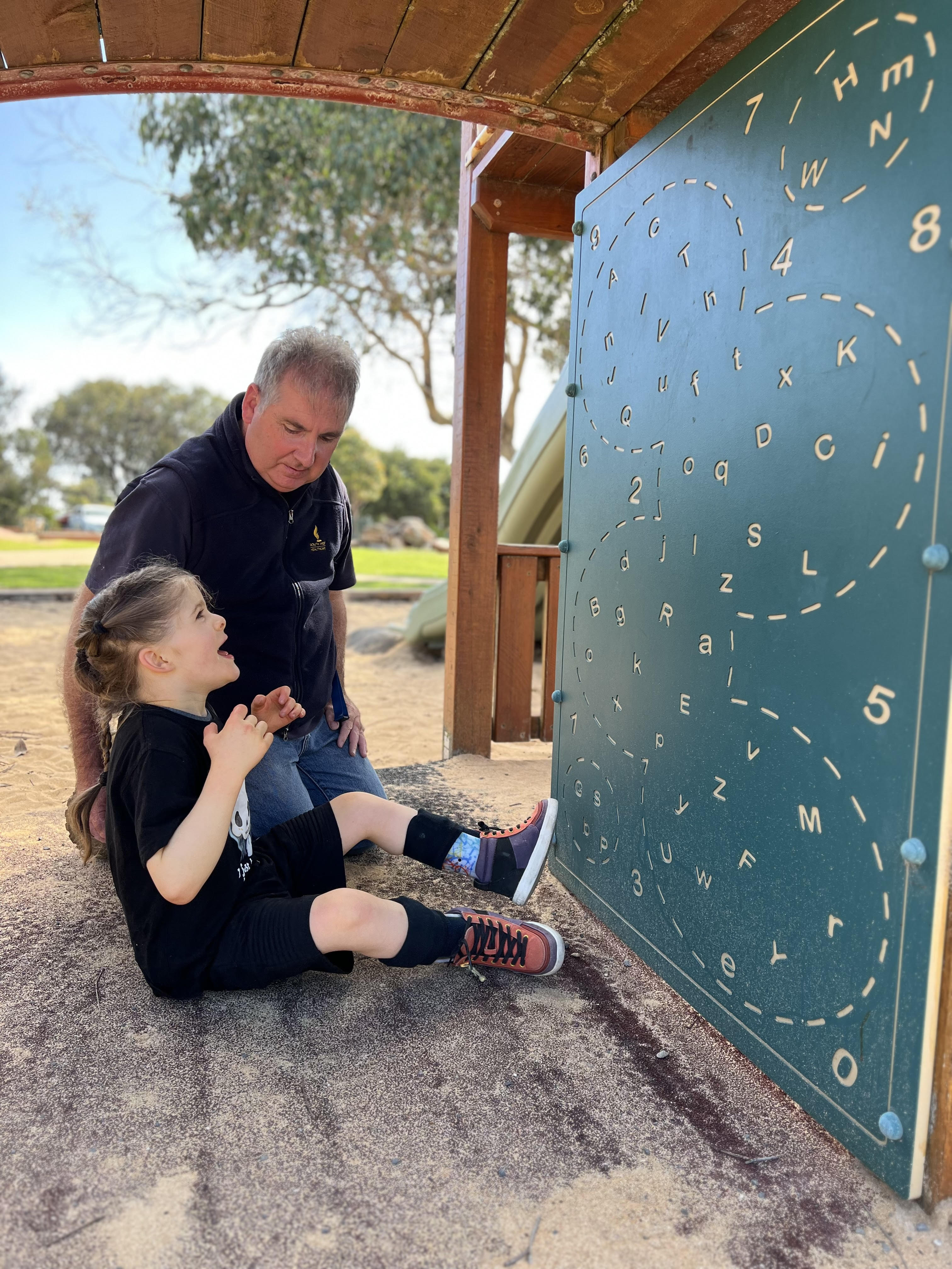 Chris and Marshall are sitting beneath wooden park play equipment. Marshall is looking at a green board with letters and numbers. He has long tied back brown hair and is wearing a black shirt and black shorts. Chris has short grey hair and is wearing a navy shirt and blue denim jeans.