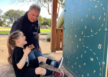 Chris and Marshall are sitting beneath wooden park play equipment. Marshall is looking at a green board with letters and numbers. He has long tied back brown hair and is wearing a black shirt and black shorts. Chris has short grey hair and is wearing a navy shirt and blue denim jeans.