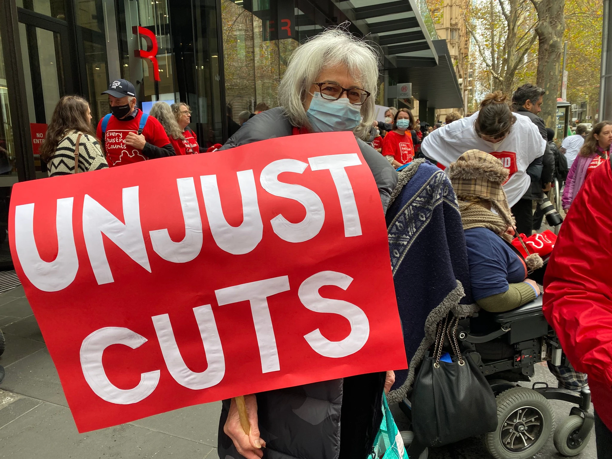 A woman in a face mask and glasses holds a large red sign with white text reading 'unjust cuts'. This woman is part of a group of advocates rallying to defend the NDIS in Melbourne as part of the Every Australian Counts campaign.