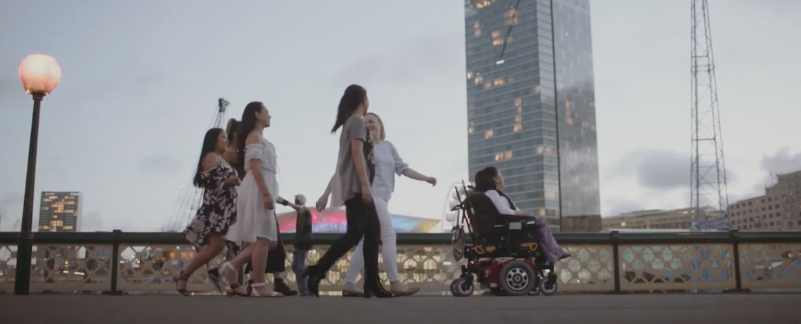 Mel and her four support workers are on a sidewalk. In the background is a city skyline at dusk. 