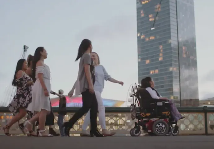 Mel and her four support workers are on a sidewalk. In the background is a city skyline at dusk.