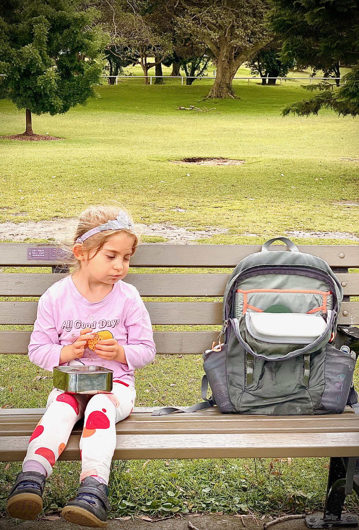 A young girl sits on a park bench eating her lunch from a silver lunch box. Her school bag sits beside her. The girl wears a floral headband, pink shirt and pink and white spotty leggings.