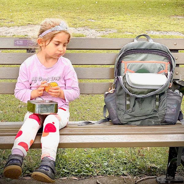 A young girl sits on a park bench eating her lunch from a silver lunch box. Her school bag sits beside her. The girl wears a floral headband, pink shirt and pink and white spotty leggings.