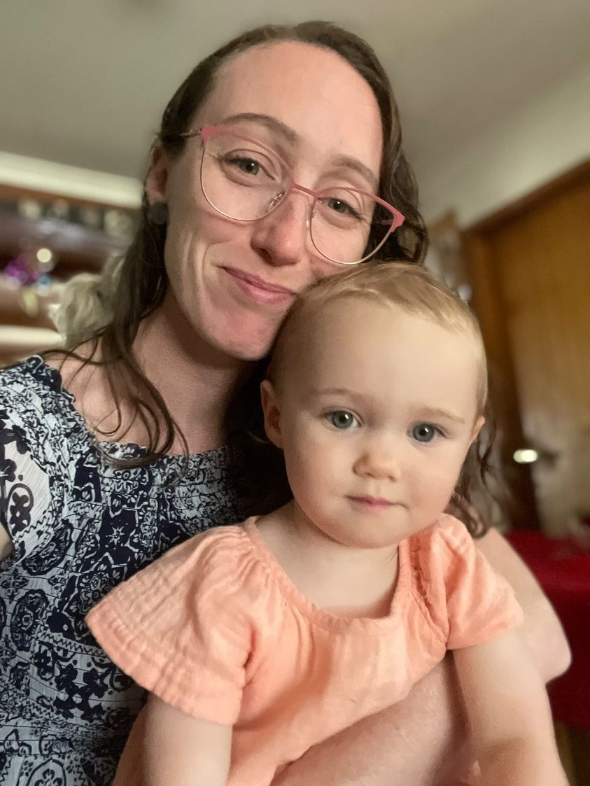 Rhi Sugars, a woman with long brown hair and glasses, smiles at the camera while holding her baby daughter. 