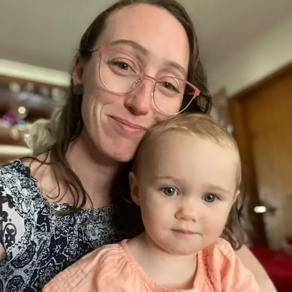 Rhi Sugars, a woman with long brown hair and glasses, smiles at the camera while holding her baby daughter.