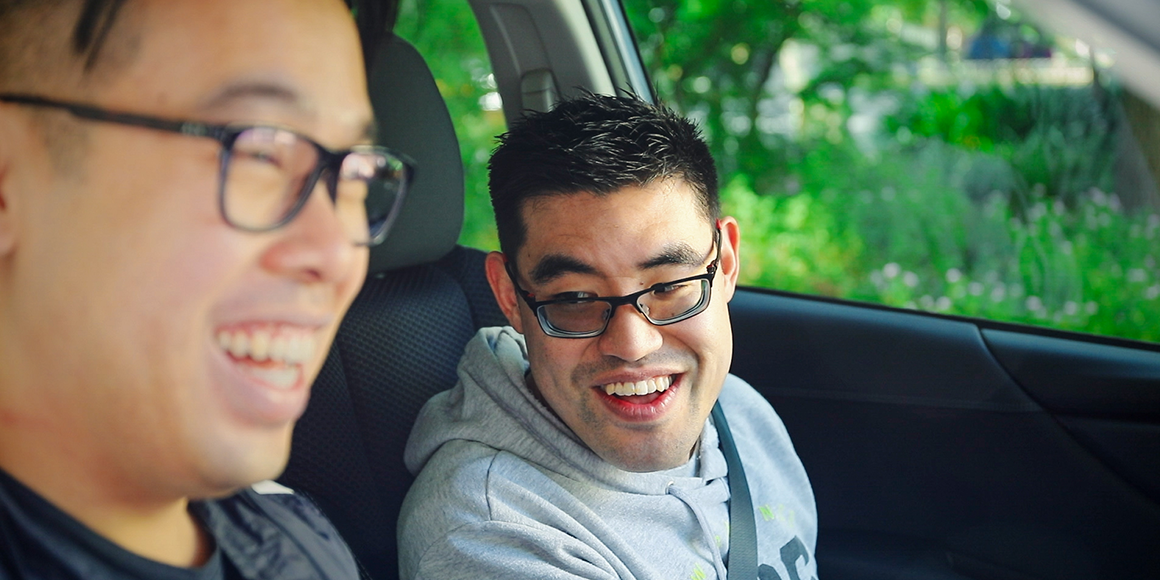 Two men with straight black hair and glasses are sitting in a car smiling. One is wearing a black t-shirt and the other is wearing a grey hoodie. They both have their seatbelts on.