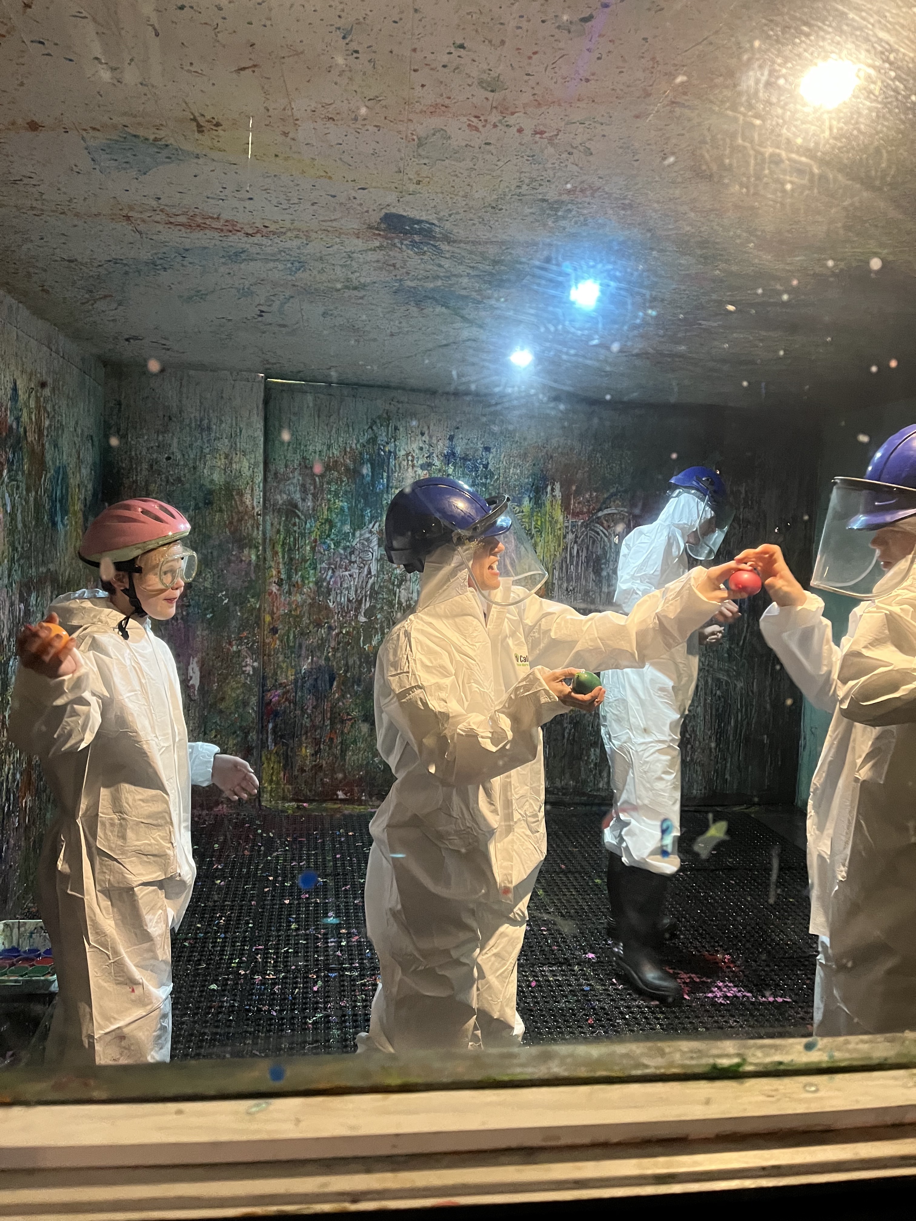 A group people wearing protective helmets and white jumpsuits are in a 'splash room' covered in paint.
