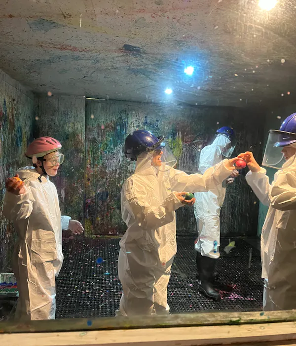 A group people wearing protective helmets and white jumpsuits are in a 'splash room' covered in paint.