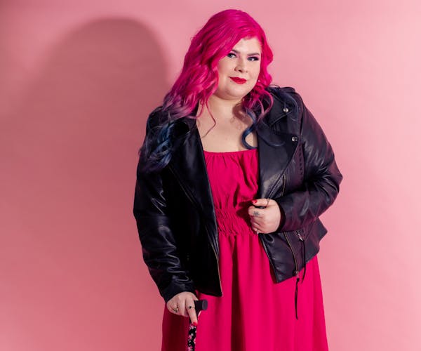 Zoe Simmons has pale skin, pink hair, and bright red lipstick. She's wearing a pink dress, black ankle boots and floral fishnets, and is sitting in a manual wheelchair adorned in fake flowers.
