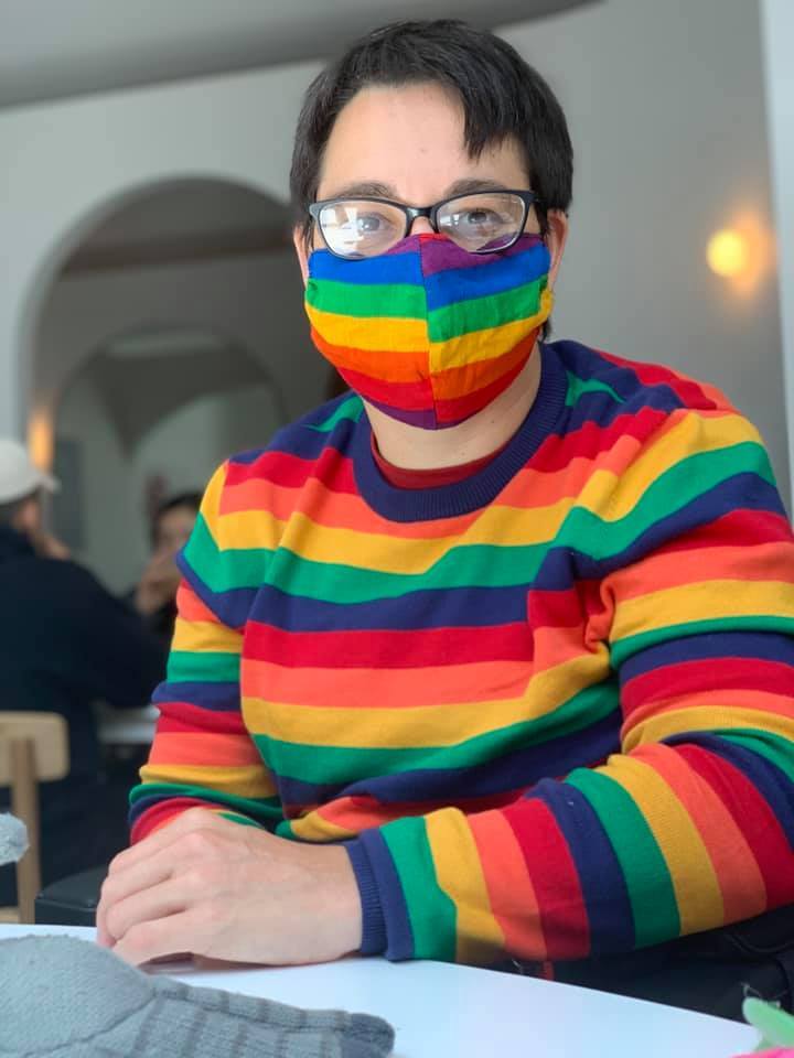 Jax has short, dark hair and is wearing a rainbow striped jumper and face mask. They also wear glasses.