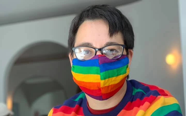 Jax has short, dark hair and is wearing a rainbow striped jumper and face mask. They also wear glasses.