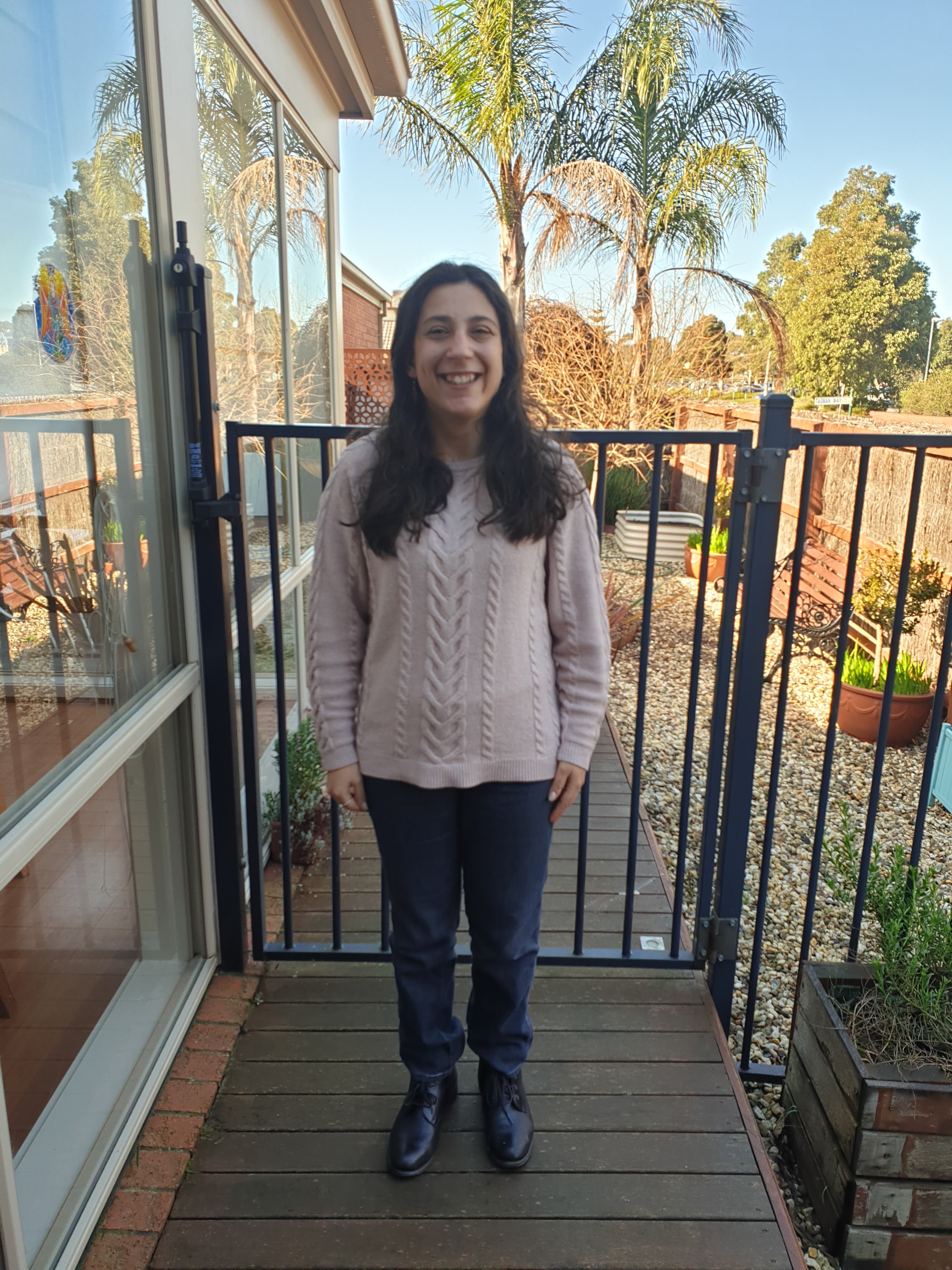 Disability activist and writer Laura Pettenuzzo stands smiling on a deck in garden on a sunny day.