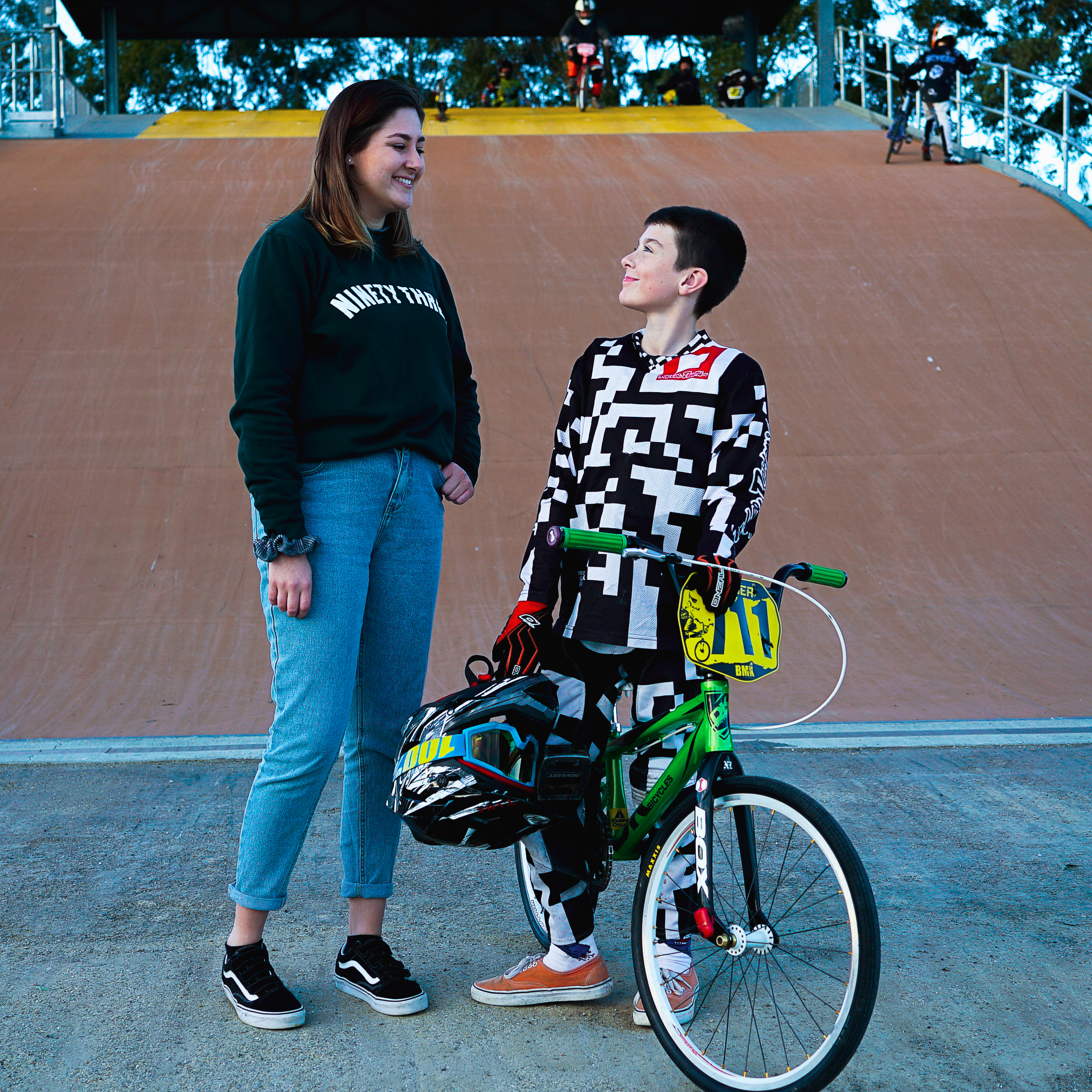 Noah is standing with his green BMX bike and looking up at Tess. Both are a smiling. Tess is wearing a black sweater and blue jeans. Noah is wearing black and white bmx gear and holding a motocross helmet and goggles. Behind them is a large brown dirt bmx start ramp.