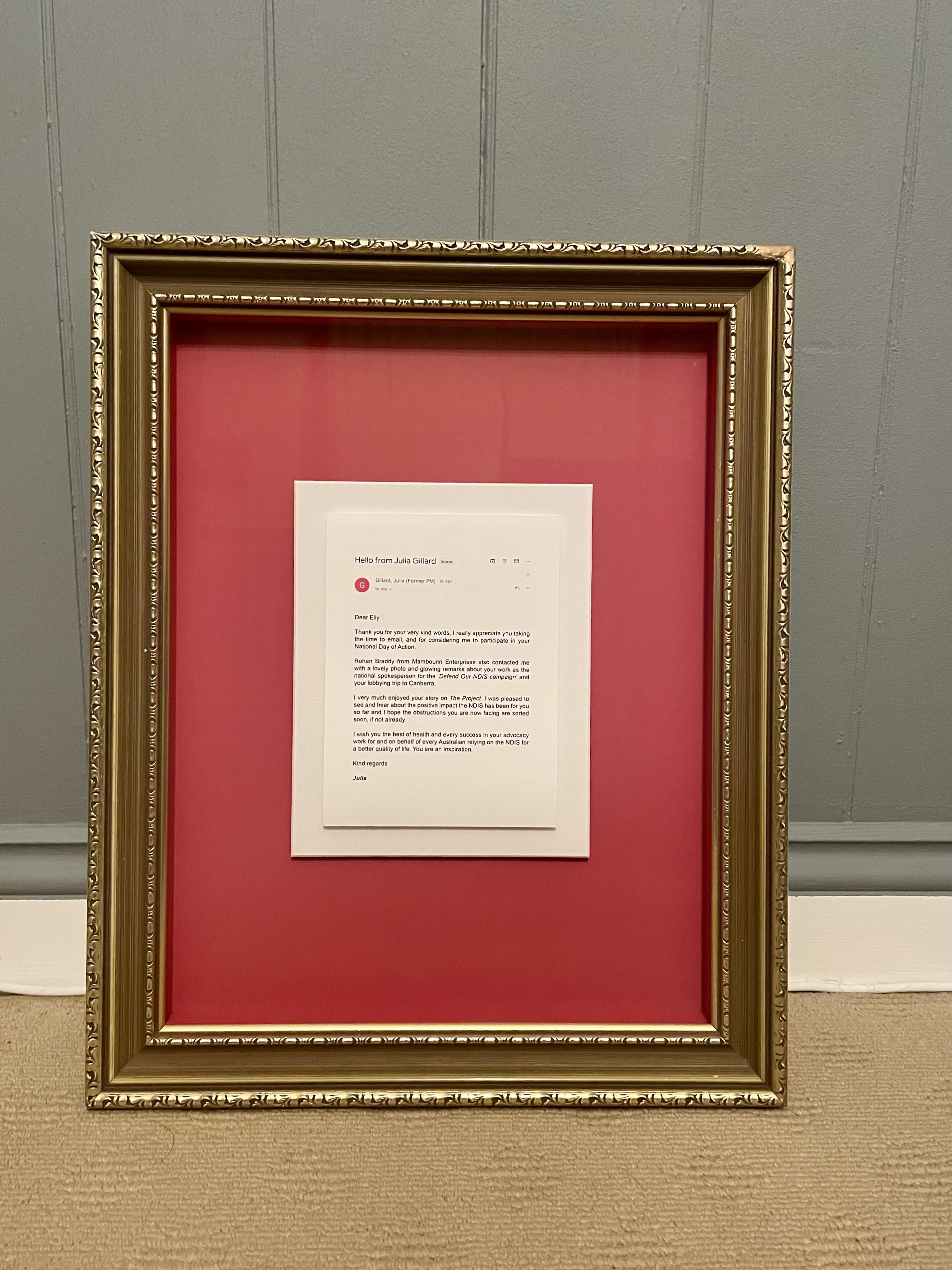An email printed out and framed. It was written by former Australian PM Julia Gillard and sent to disability advocate Elly Desmarchelier. The email is printed on cream paper and set against a red background. The picture frame is ornate and gold. 
