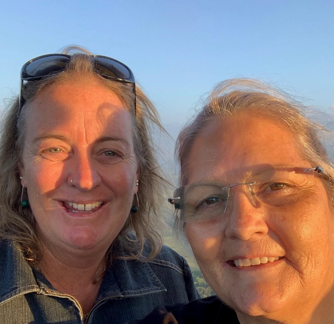 Michelle has grey hair tied back and is wearing clear spectacles. Annette has blonde hair being held back by sunglasses resting on her head. The sun is shinning on their faces.