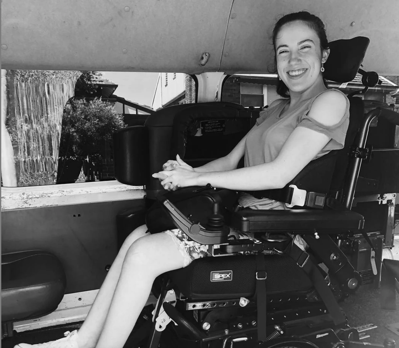 Hannah is smiling toward the camera. She is using her wheelchair and is in the back of her van.
