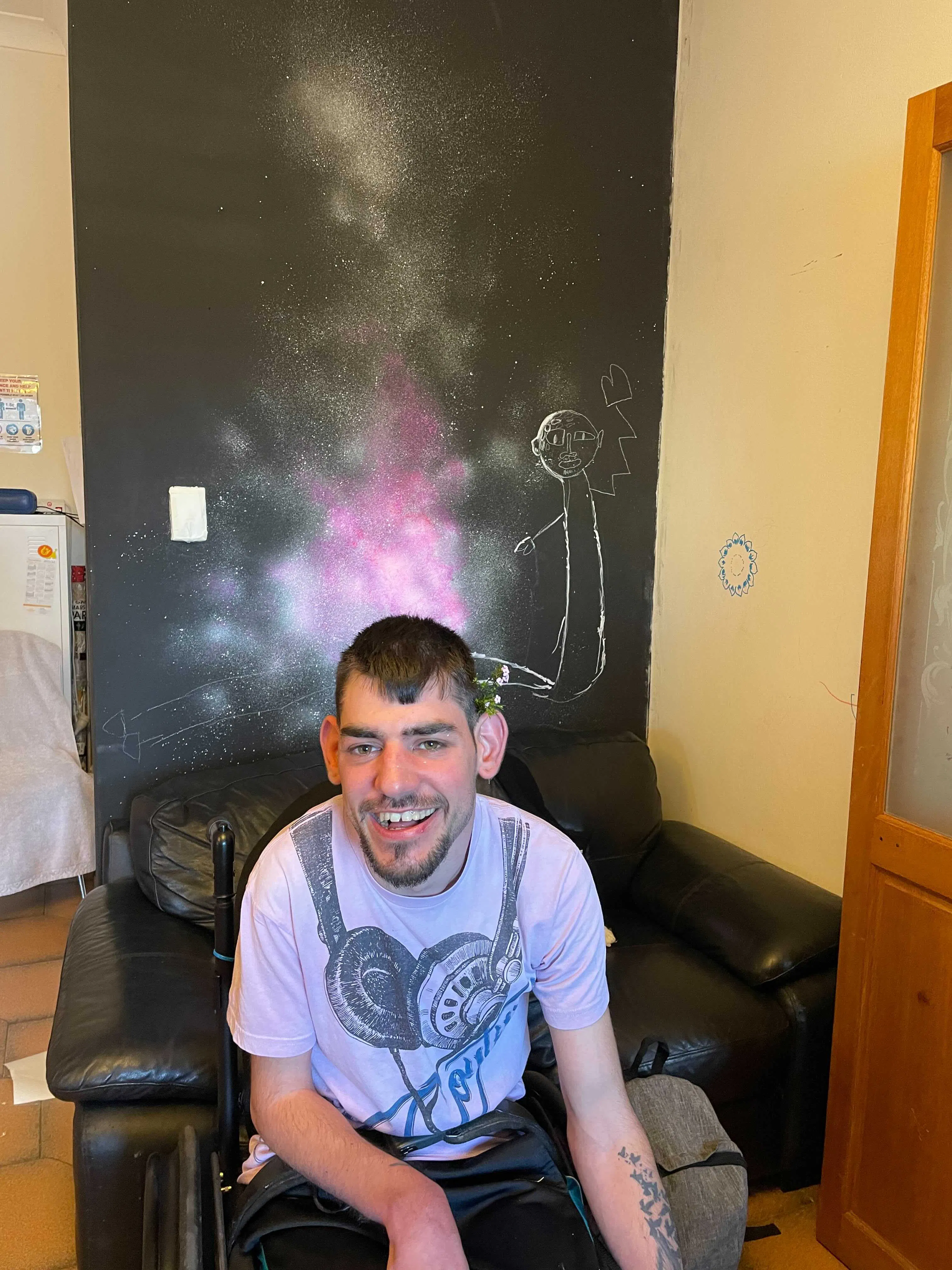 Andrew is sitting in front of the mural in progress. He is smiling. Andrew uses a wheelchair. His shirt has a picture of headphones.