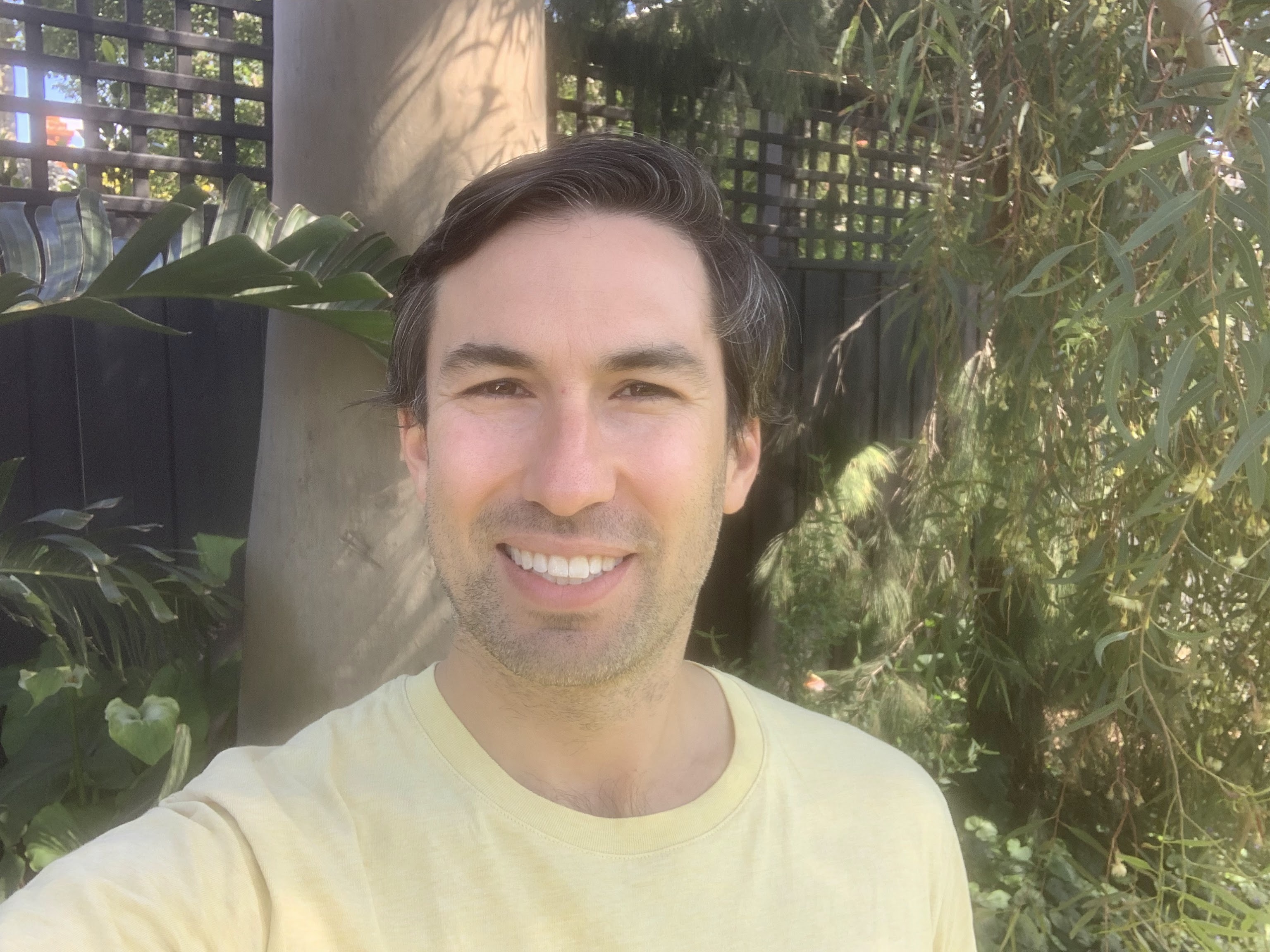 Joe is smilig to camera. Joe has short dark hair and some stubble facial hair. Behind him are trees and a fence.