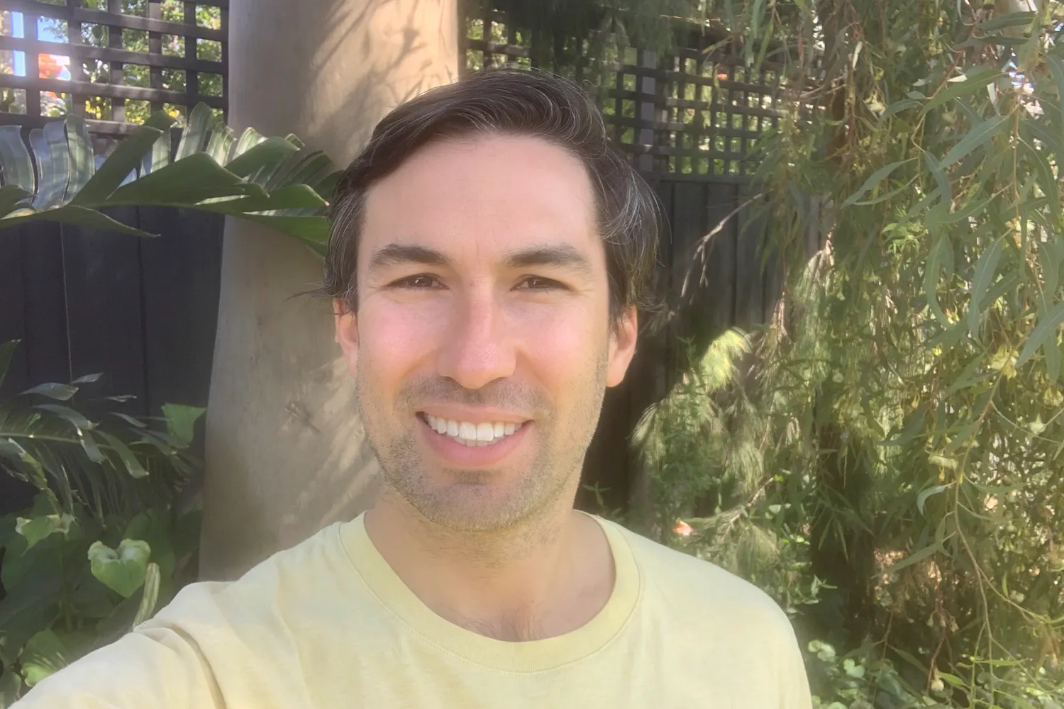 Joe is smilig to camera. Joe has short dark hair and some stubble facial hair. Behind him are trees and a fence.
