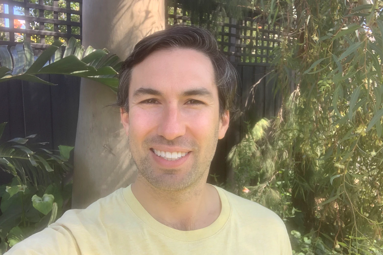 Joe is smilig to camera. Joe has short dark hair and some stubble facial hair. Behind him are trees and a fence.