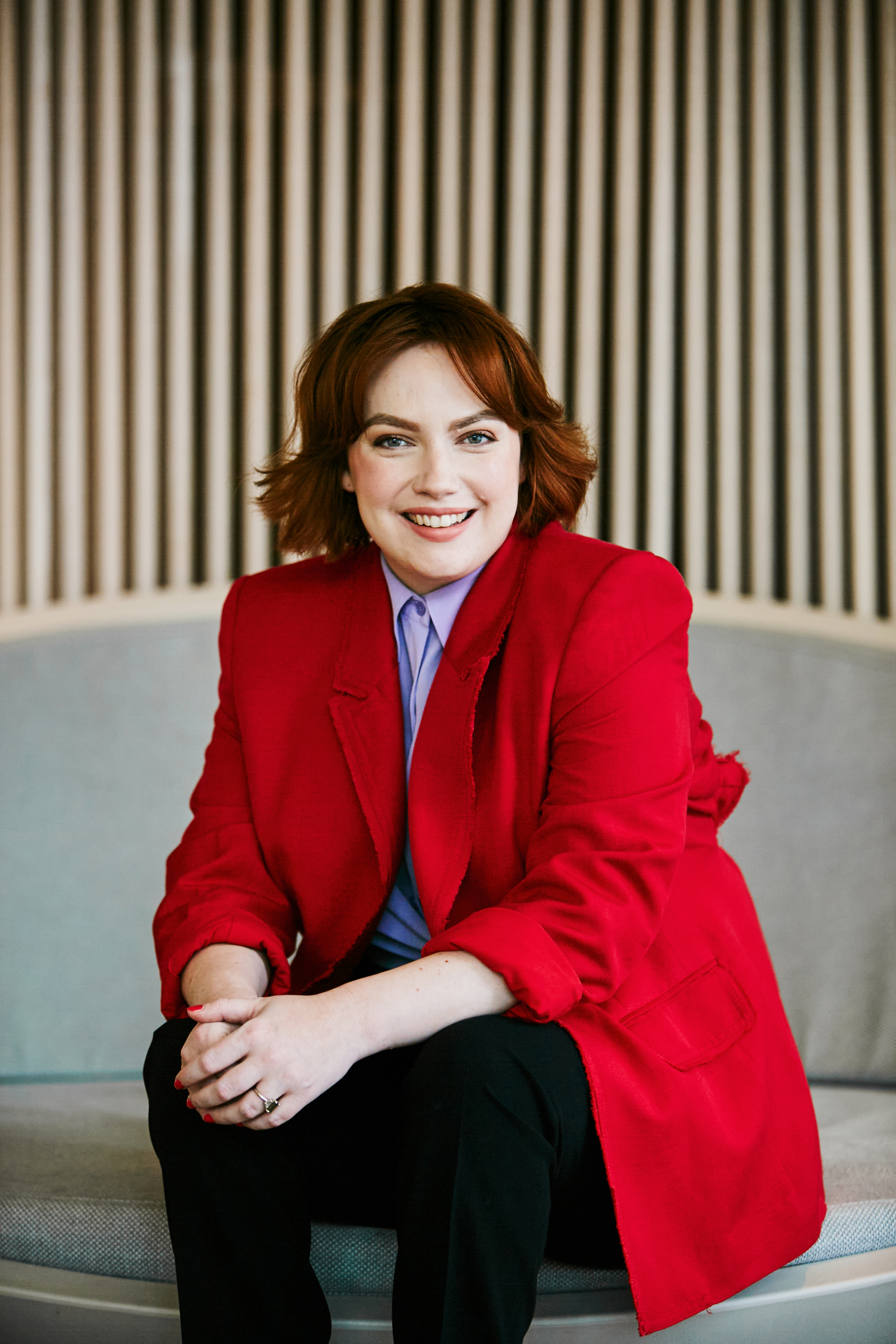 Disability rights advocate Elly Desmarchelier has shoulder-length red hair and wears a red blazer and purple shirt. She smiles at the camera and poses in a relaxed seated position.