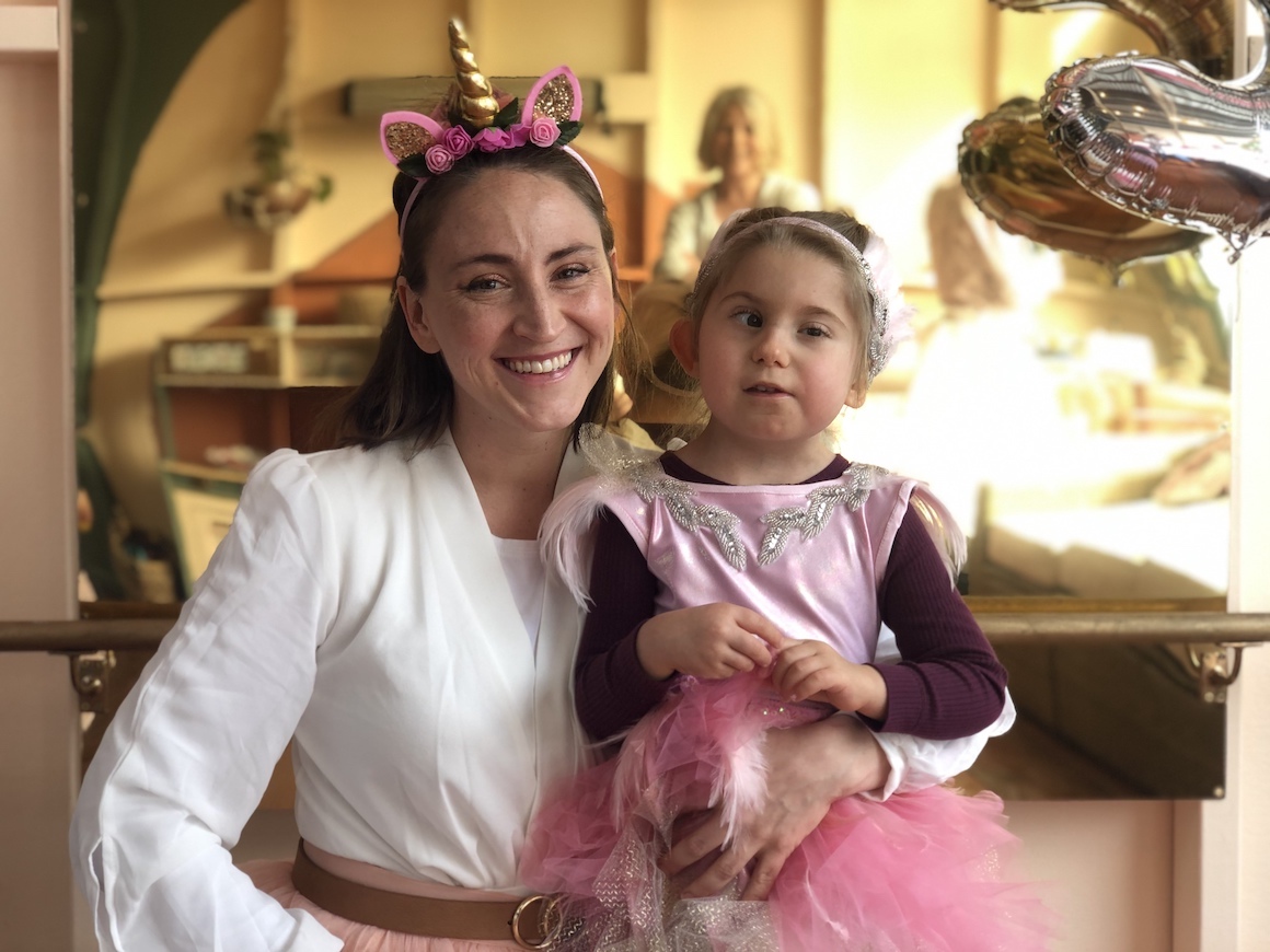 Holly is wearing a unicorn headband and is smiling. Her arm is wrapped around her daughter who's wearing a pink tutu. 