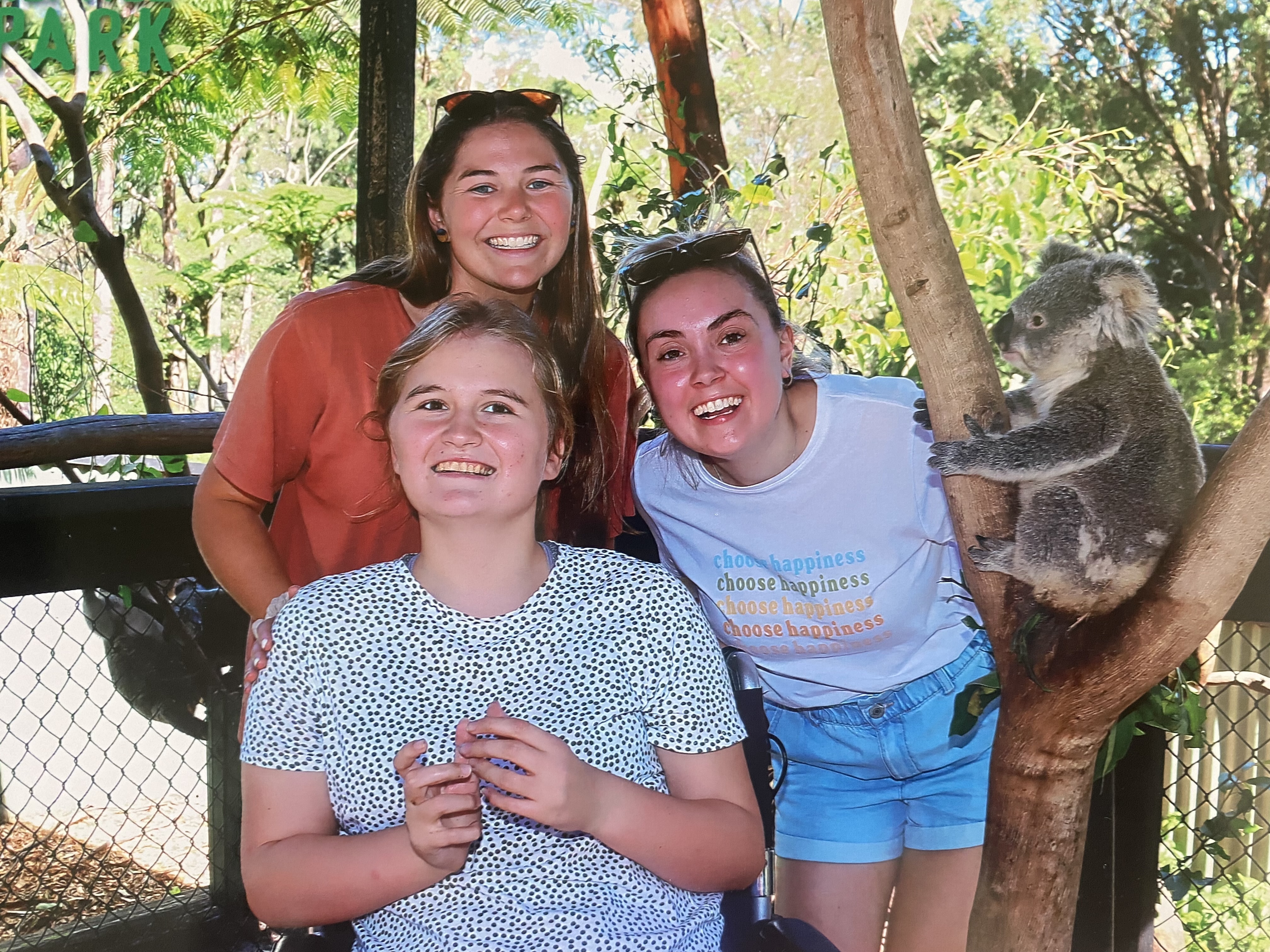 Abbey and her two support workers are smiling. They're at a koala sanctuary, and are next to a tree with a koala resting on the branches. Abbey is a wheelchair user.
