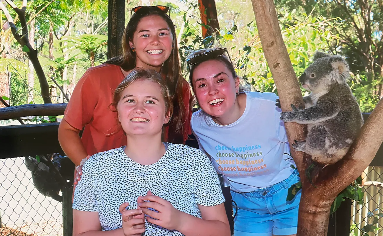 Abbey and her two support workers are smiling. They're at a koala sanctuary, and are next to a tree with a koala resting on the branches. Abbey is a wheelchair user.
