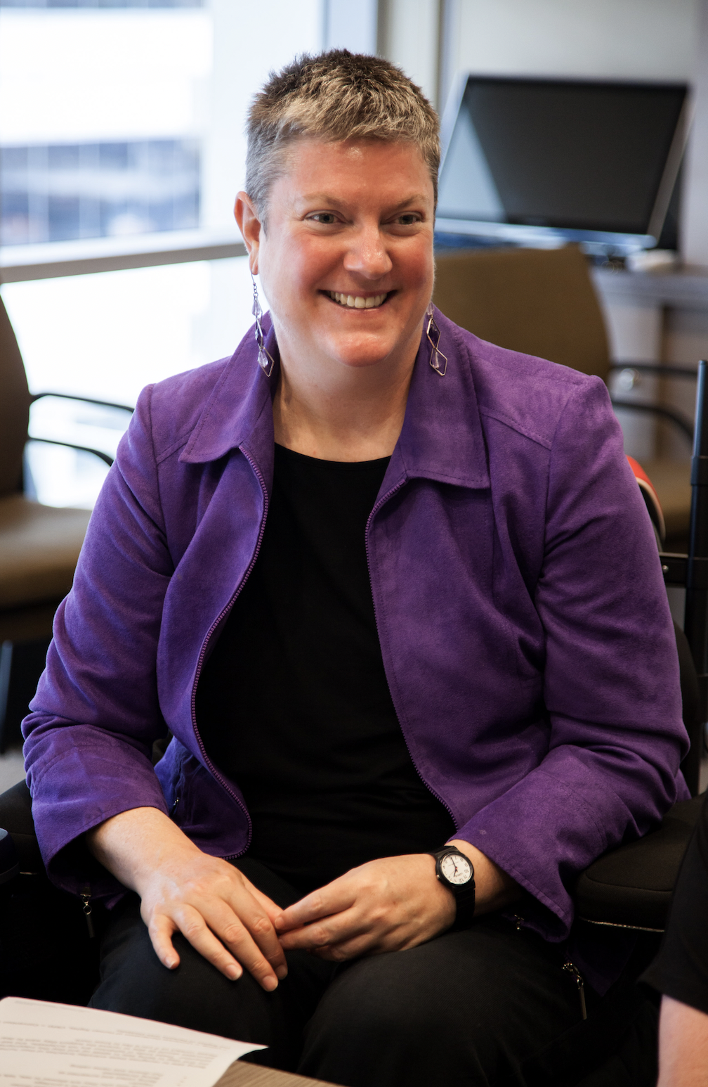 Image shows: Christina Ryan seated and smiling at someone (off camera). She is wearing a purple blazer over a black outfit and has short grey hair. 