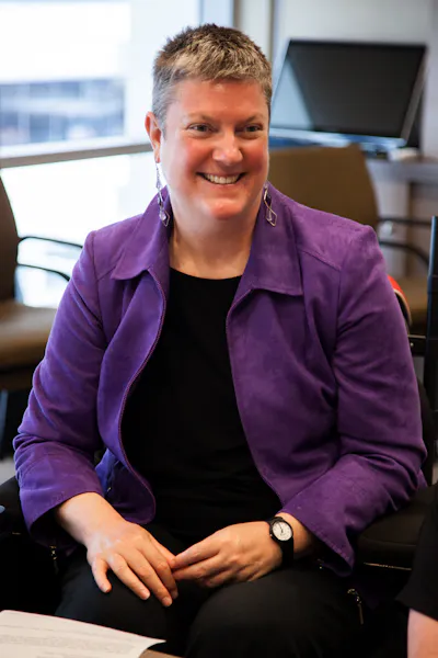 Image shows: Christina Ryan seated and smiling at someone (off camera). She is wearing a purple blazer over a black outfit and has short grey hair.