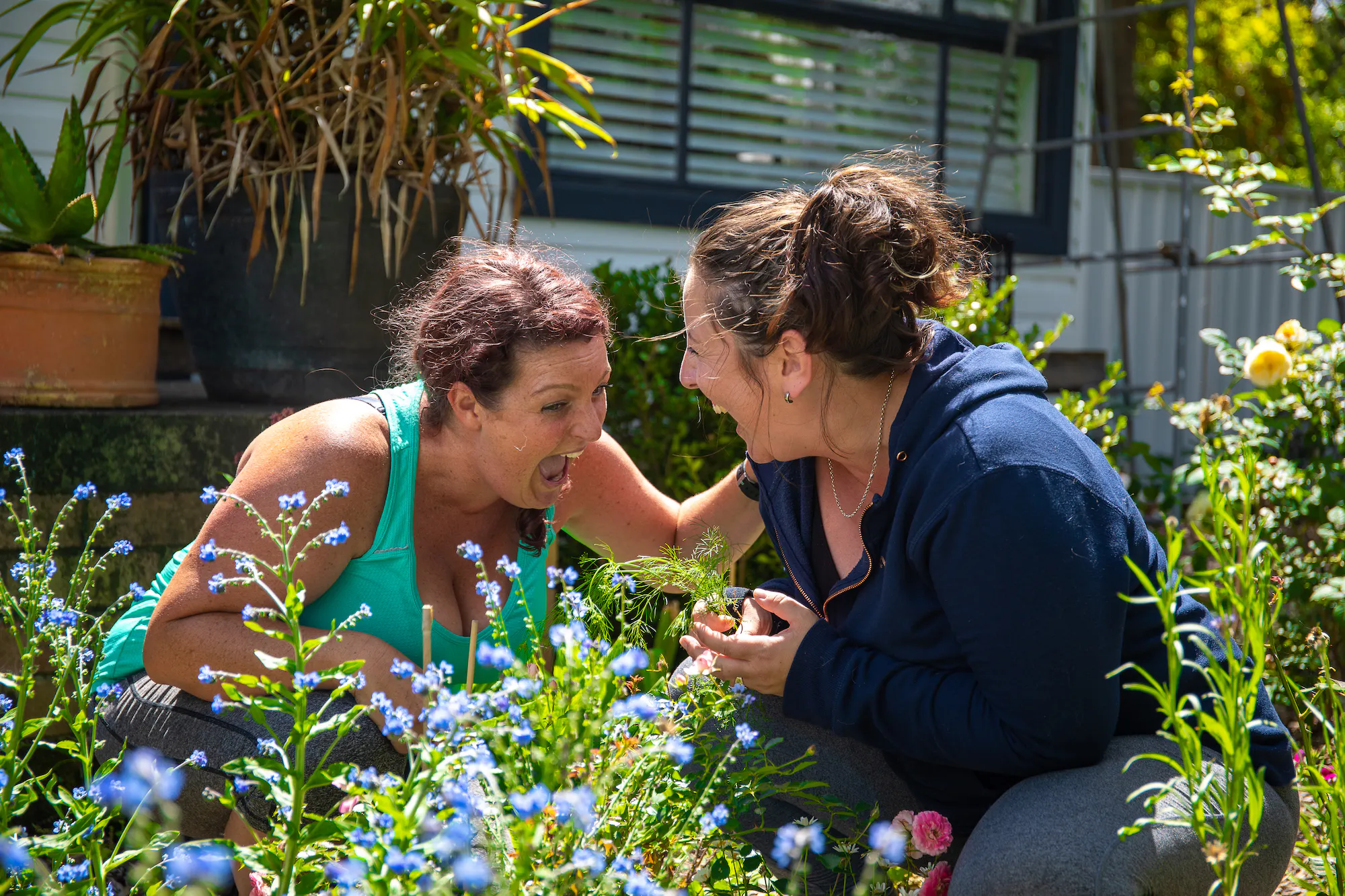 Portia and Sam are knelt down in the garden with flowers all around them. They are staring at each other, laughing uncontrollably. 