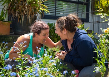 Portia and Sam are knelt down in the garden with flowers all around them. They are staring at each other, laughing uncontrollably.