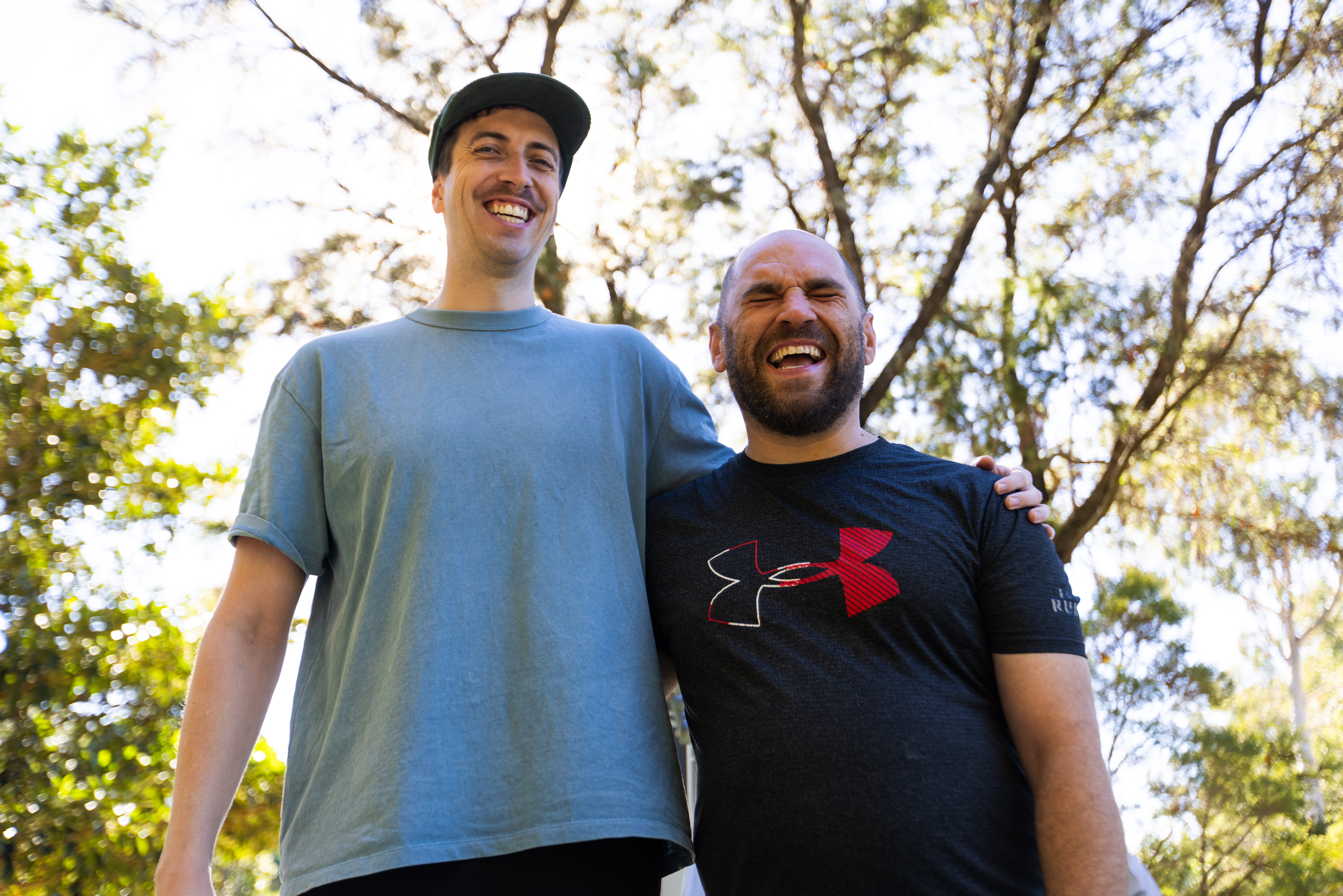 Tony and his Hireup support worker David are standing side by side in a park, with an arm wrapped tightly across each other's shoulders. Sunlight seeps through the leafy trees. Tony is bald, has a beard and wears a navy top. David has a moustache, wears a cap and light blue top. 